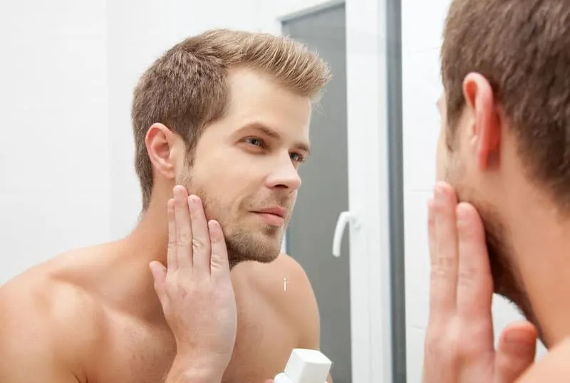 man touching his face infront of the mirror wearing no shirt 