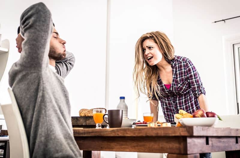 nagging woman hard in the morning in front of a man sitting in the table with breakfast food