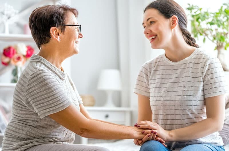 mulher mais velha toca na mão da mulher mais nova colocada no seu colo enquanto está sentada na sala de estar