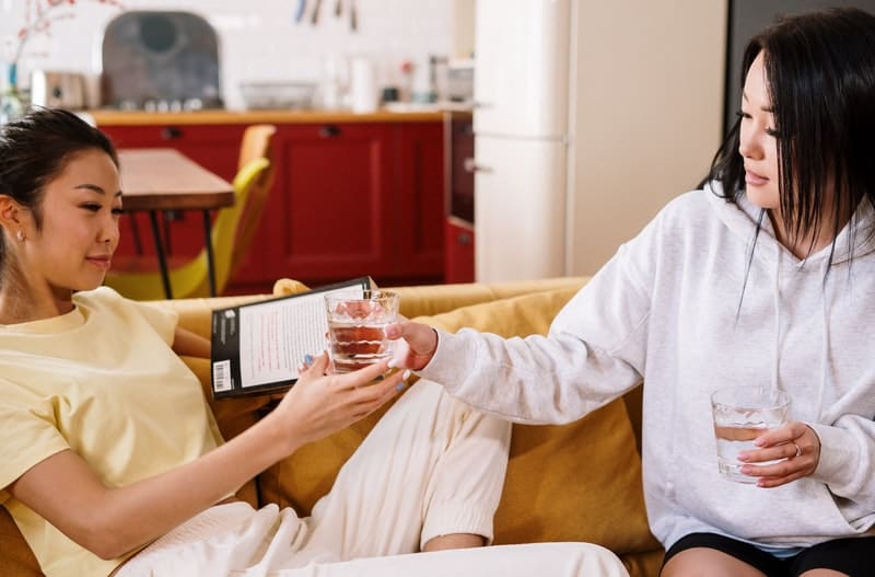 person holding clear glass giving it to another woman holding a tab sitting beside her in the sofa