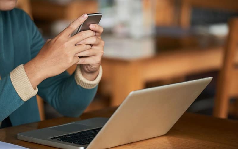 person holding smartphone with both hands in front of a laptop on a table