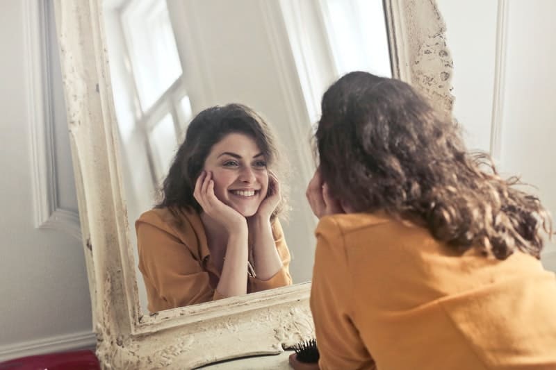 foto di donna rivolta allo specchio che sorride con le mani che sostengono la testa