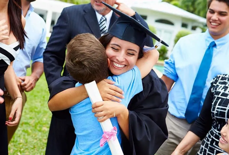 proud little brother hugging woman in graduation gown with people around them