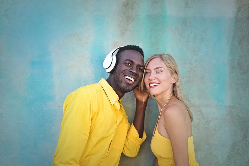 smiling man and woman standing near blue wall listening music from headphones