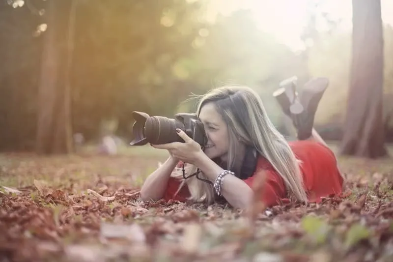 smiling woman taking photos on professional camera on dried leaves on the ground