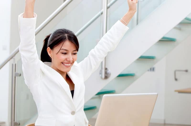 successful woman raising her hands in front of a laptop in an office set up near the stairs