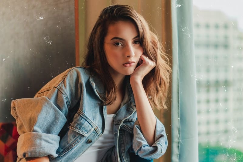 thinking woman in denim jacket with hand supporting her head while sitting near windows