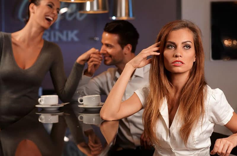 unhappy woman sitting on the bar with a man sitting near him flirting on another woman