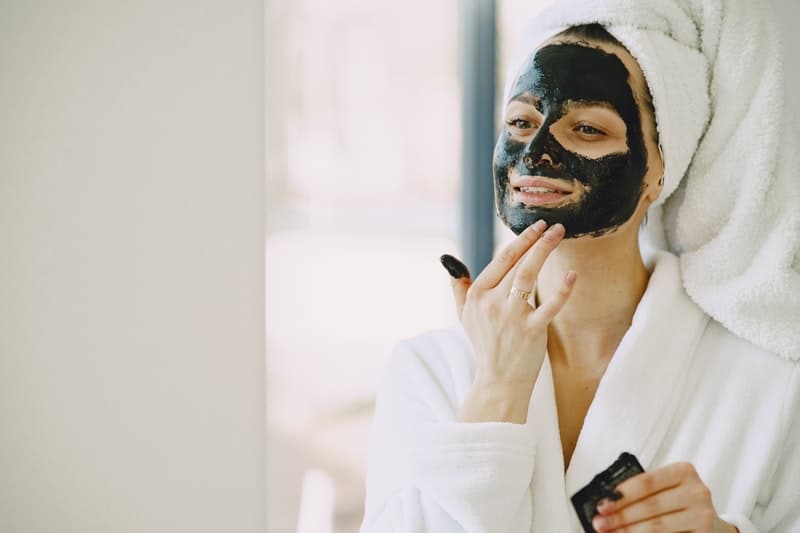 donna che applica una maschera facciale sul viso colorata di nero e indossa un accappatoio e un asciugamano sulla testa