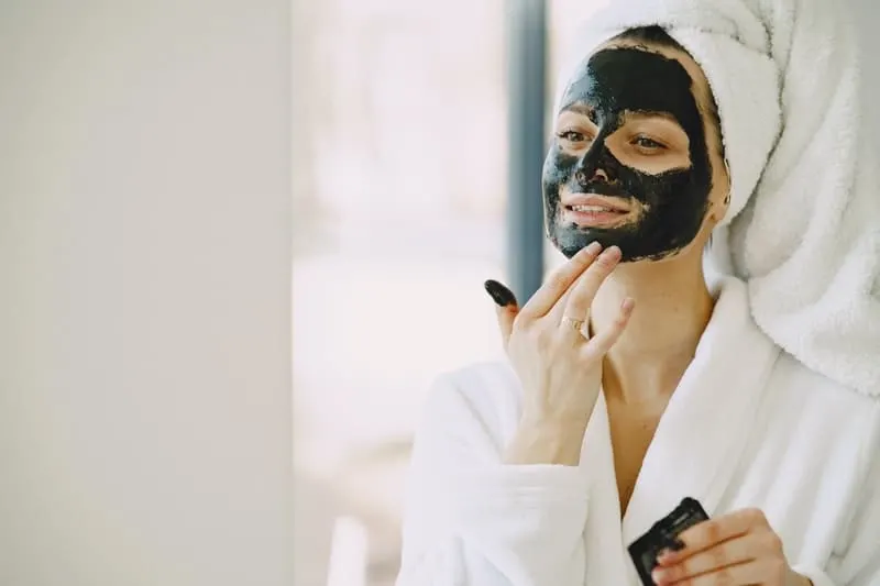 woman applying facial mask on her face colored black and wearing a bath robe and towel on her head
