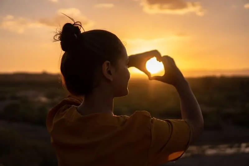 woman doing hand heart sign with the setting sun captured inside the hand sign