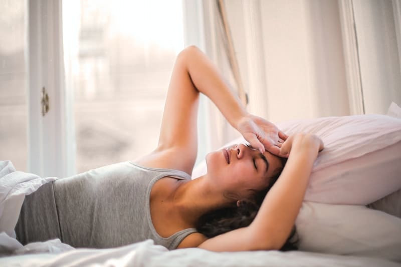 woman in gray tank top lying on bed with hand on her forehead