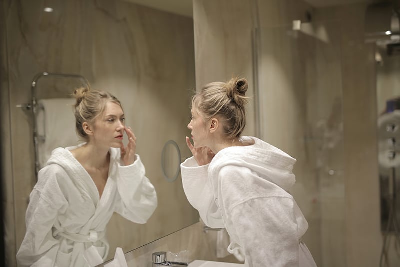 mujer en bata blanca frente al espejo tocandose la cara