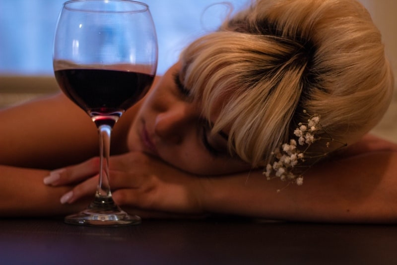 donna che appoggia la testa sul tavolo accanto al bicchiere di vino mezzo pieno