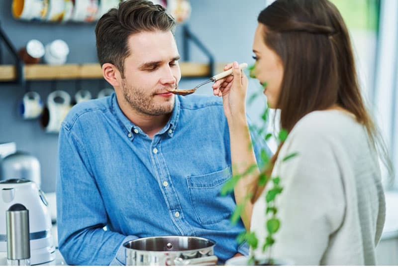 woman letting the man taste the food thru the spoon while man close his eyes tasting inside kitchen