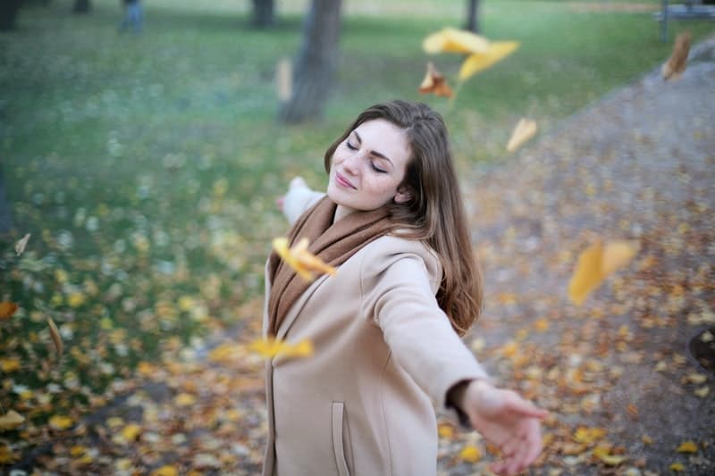 woman open arms while smiling and closing her eyes with falling dried leaves around