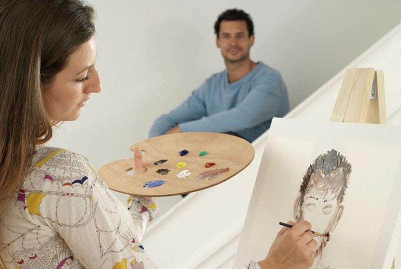 mulher a pintar o retrato de um homem sentado num lugar distante