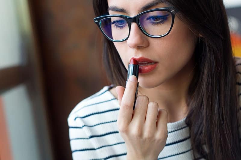 woman putting on lipstick wearing an eyeglass and striped top