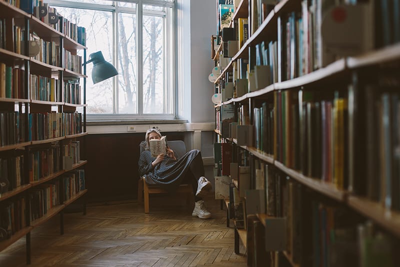 mulher a ler um livro sentada numa cadeira numa biblioteca