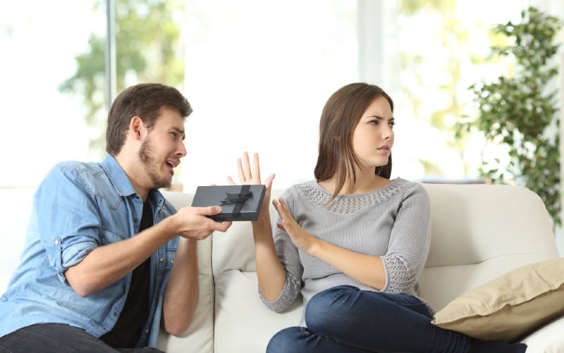 Angry woman sitting on couch rejecting gift from man