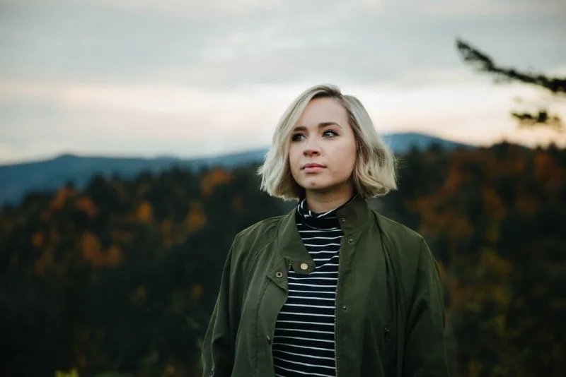 blonde woman in green jacket standing in forest