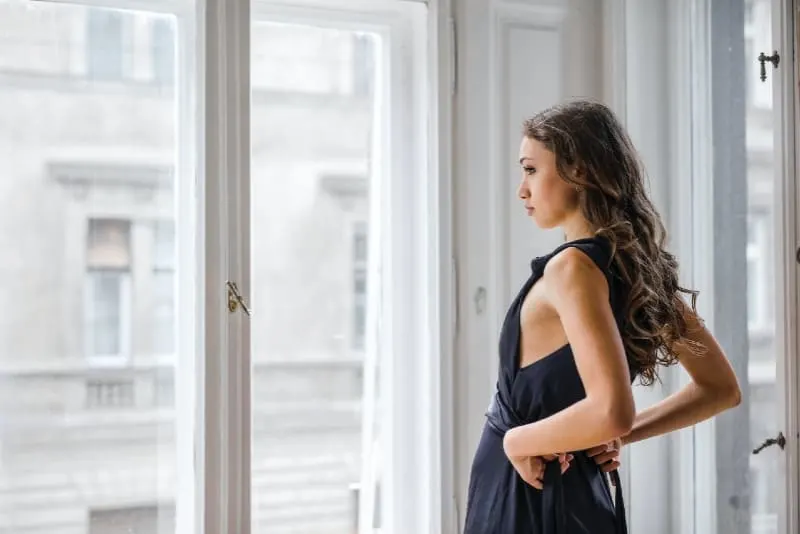 woman in black dress standing near window looking outside