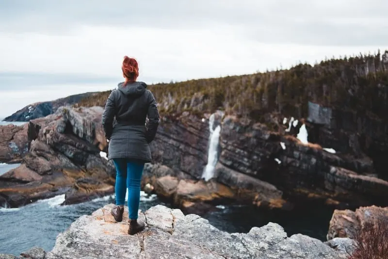 woman in black jacket standing on cliff looking at water