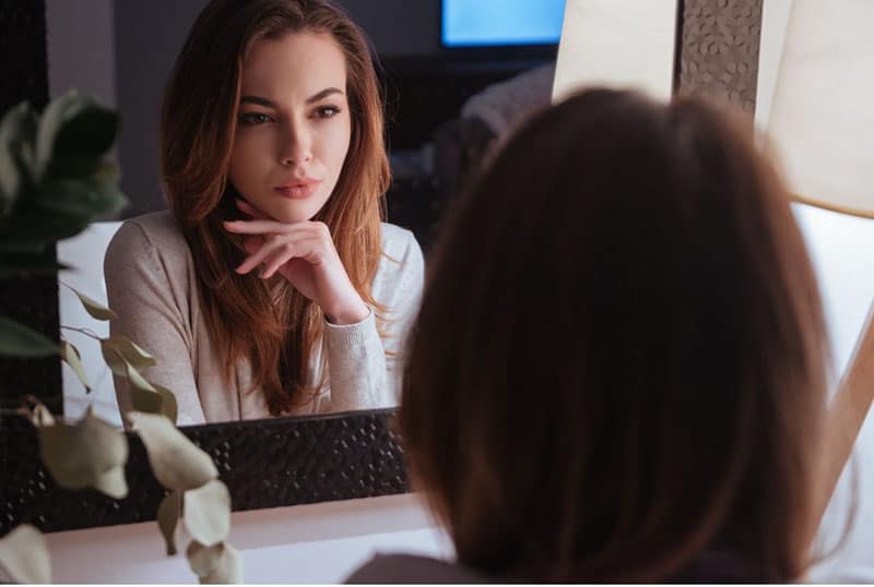 woman stares at the mirror with her image with hand on her chin 