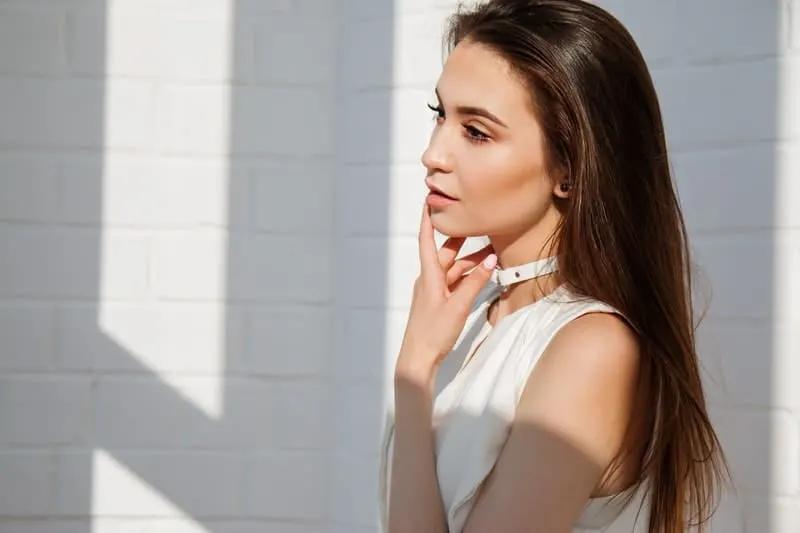 woman thinking wearing white top near white wall with hand placed near her face