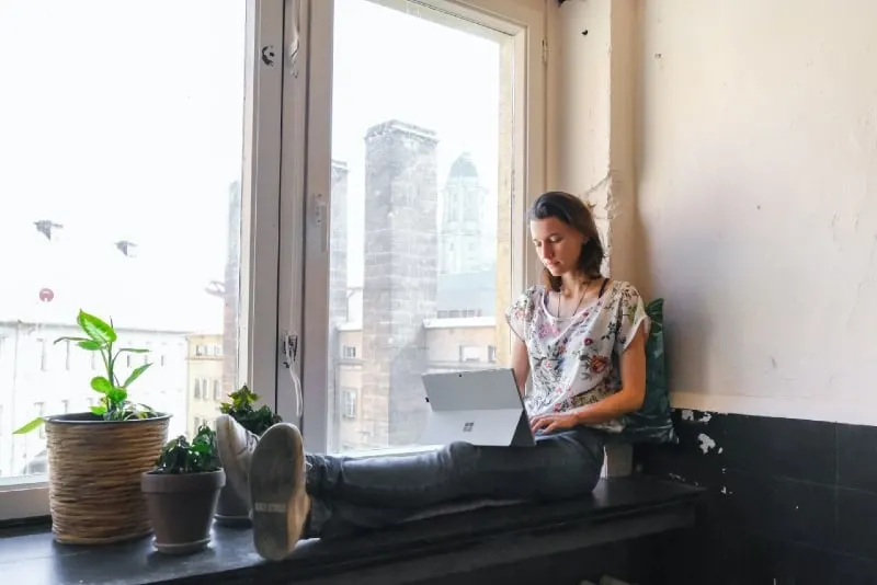 woman using laptop while sitting on window pane
