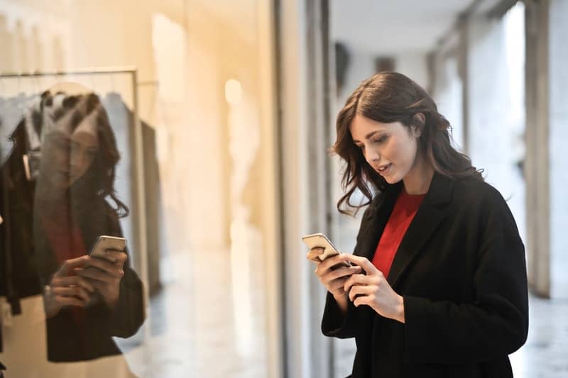 woman wearing black coat looking at her smartphone near a boutique inside a mall