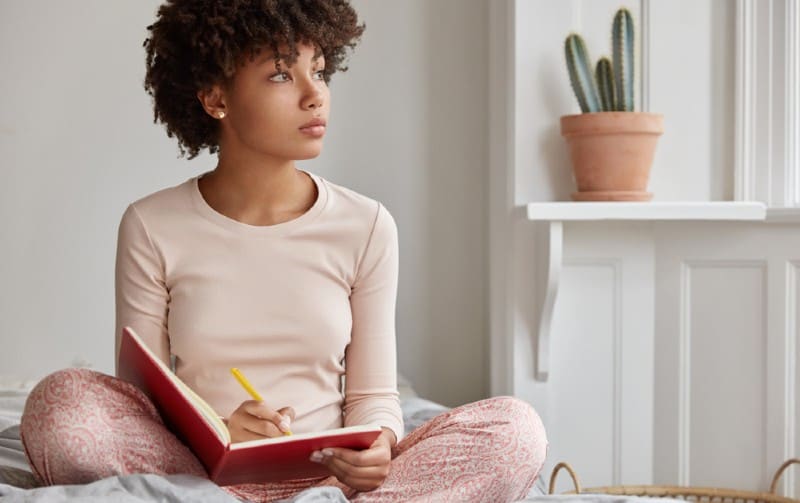 Mujer afroamericana escribiendo en un cuaderno rojo sentada en una cama