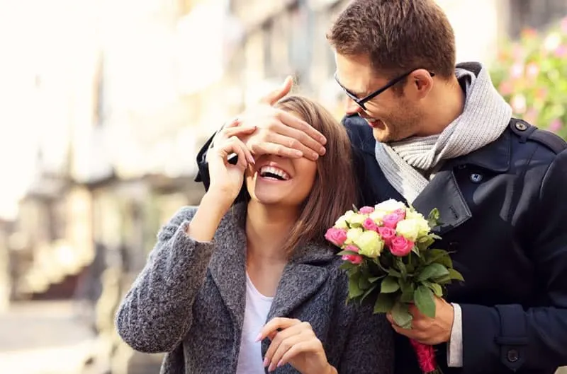 young man surprising woman with flowers and covering her eyes with his hand