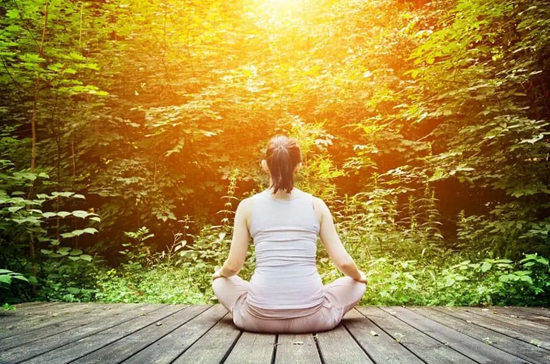 young woman meditates in front of the forest while sitting on wooden floor