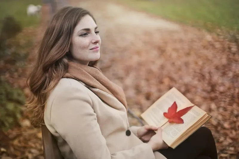 young woman with book in an autumn park sitting on a bench smiling