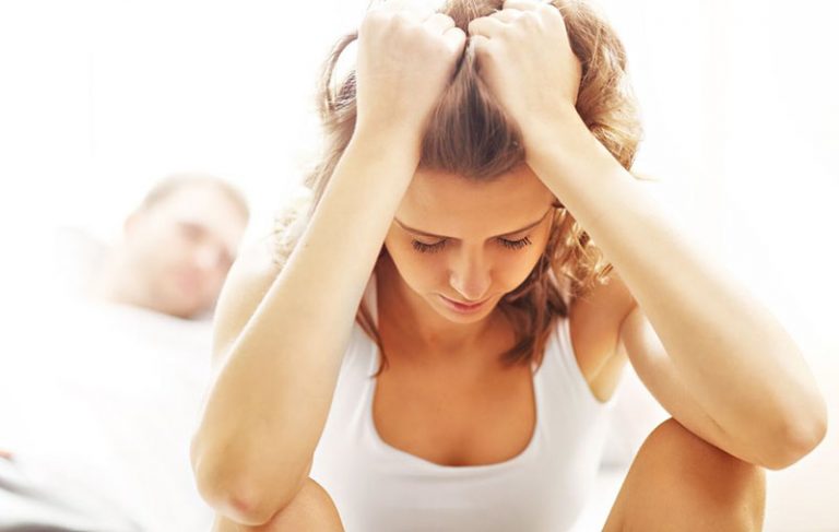 mujer apenada con las manos agarrándose el pelo mientras está sentada en la cama donde yace un hombre