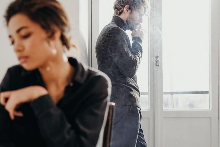 man standing while smoking near the window distant to a woman sitting