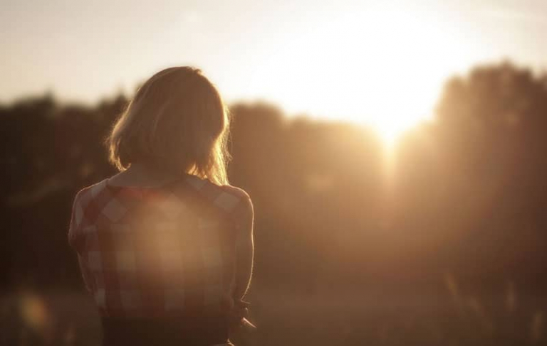 woman standing in the field facing the sunset covered with trees