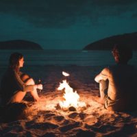 man and woman sitting on seashore near bone fire