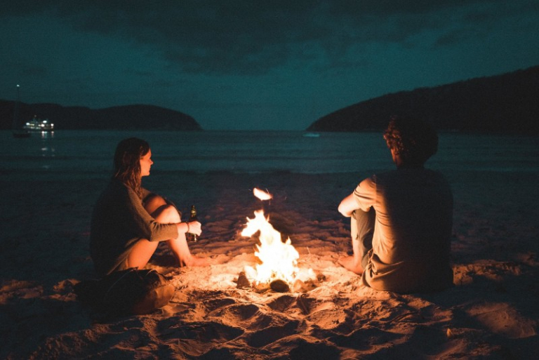 man and woman sitting on seashore near bone fire