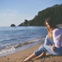 pensive woman sitting in the sand along the shore wearing white top and blue pants