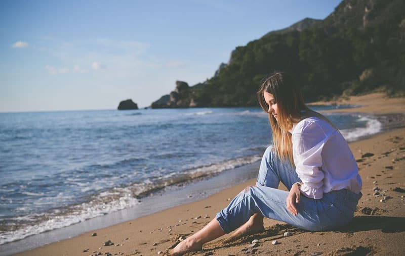 pensive woman sitting in the sand along the shore wearing white top and blue pants