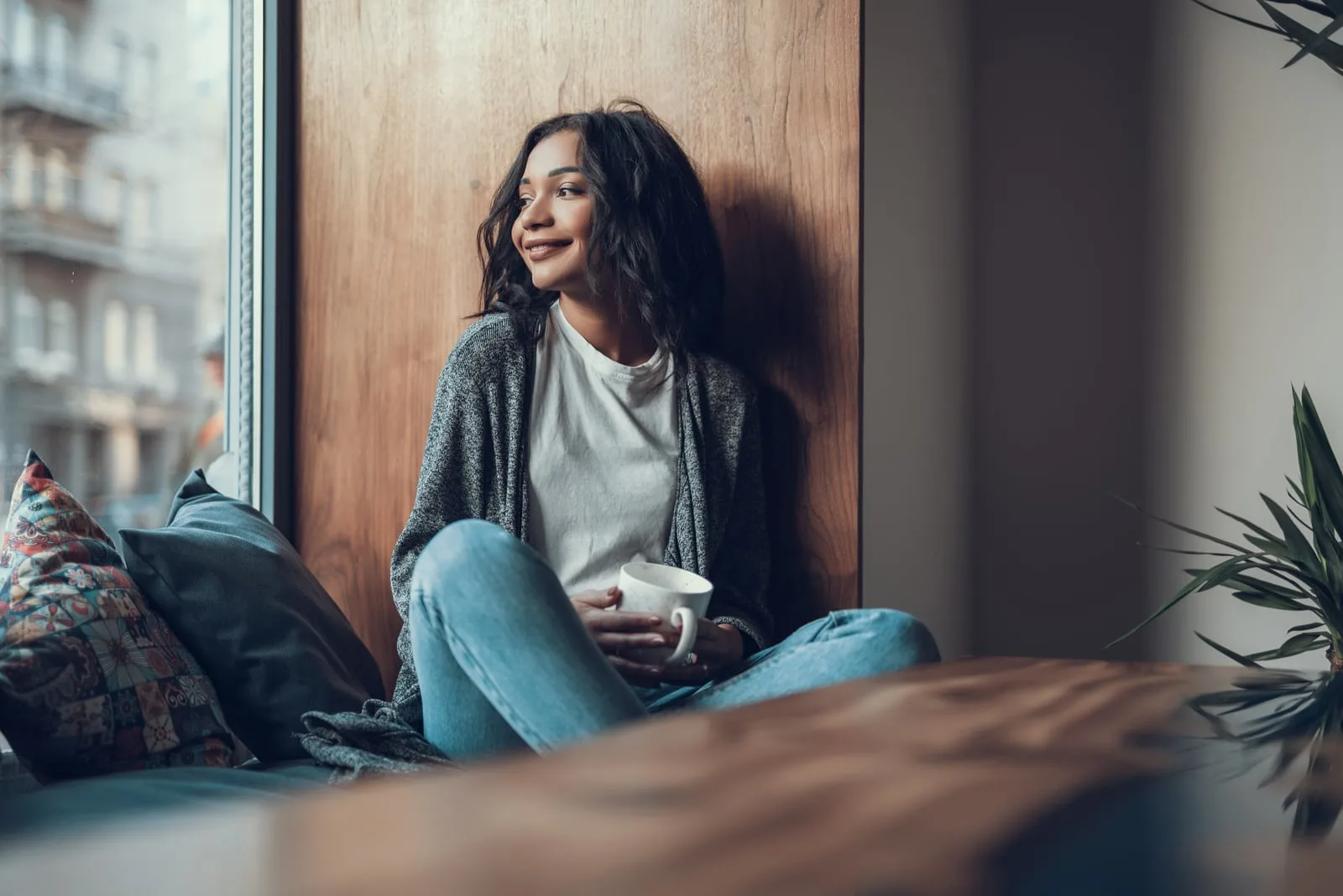 a smiling black woman sits drinking coffee and looking out the window