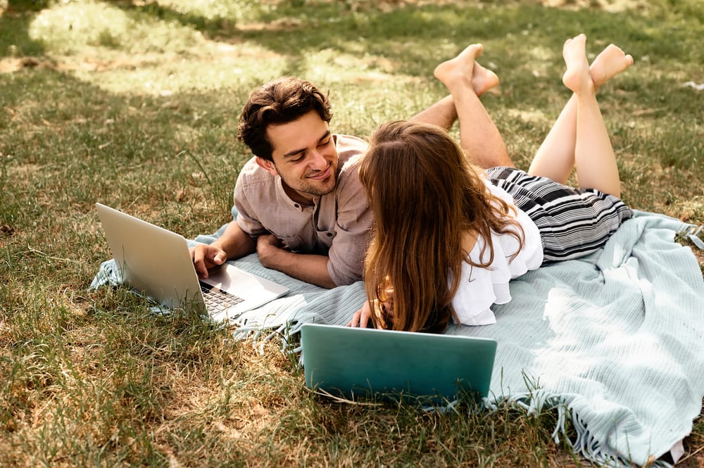 una giovane coppia di innamorati sdraiata su coperte sull'erba e che usa un computer portatile