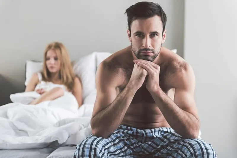 abused young man sitting on bed with wife at the back thinking