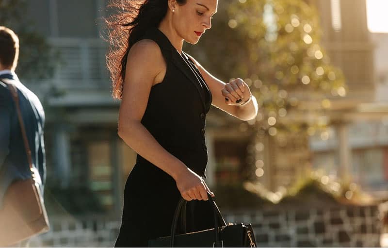 Donna d'affari che controlla l'ora sull'orologio da polso e porta con sé una borsa indossando un abito nero
