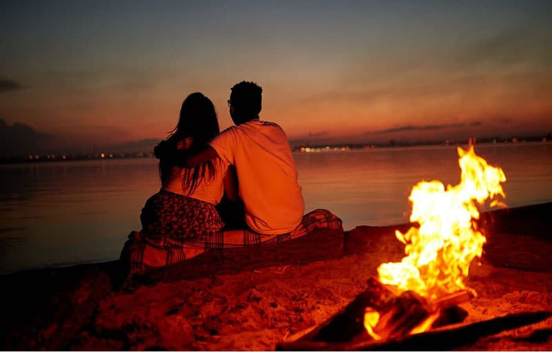 casal a namorar na praia, de frente para o mar, com uma fogueira nas costas