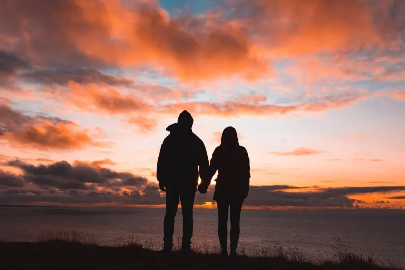 man and woman holding hands while standing on cliff during sunset