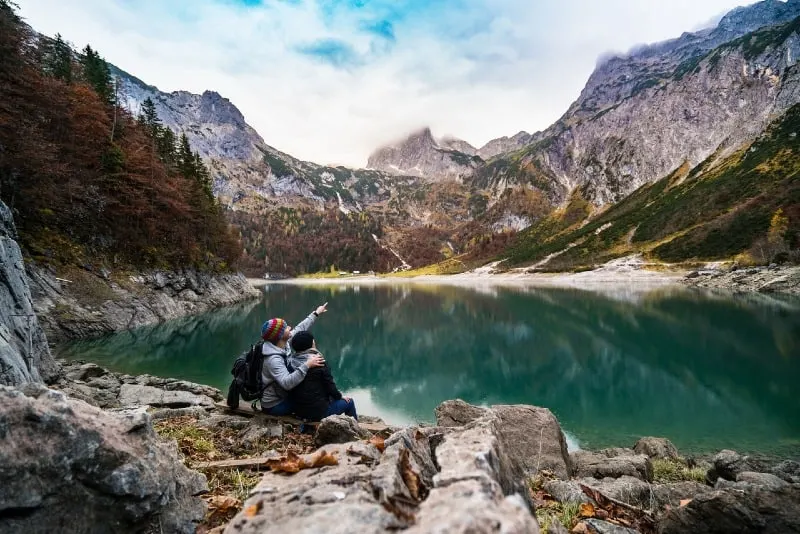 man and woman sitting near lake looking at mountain