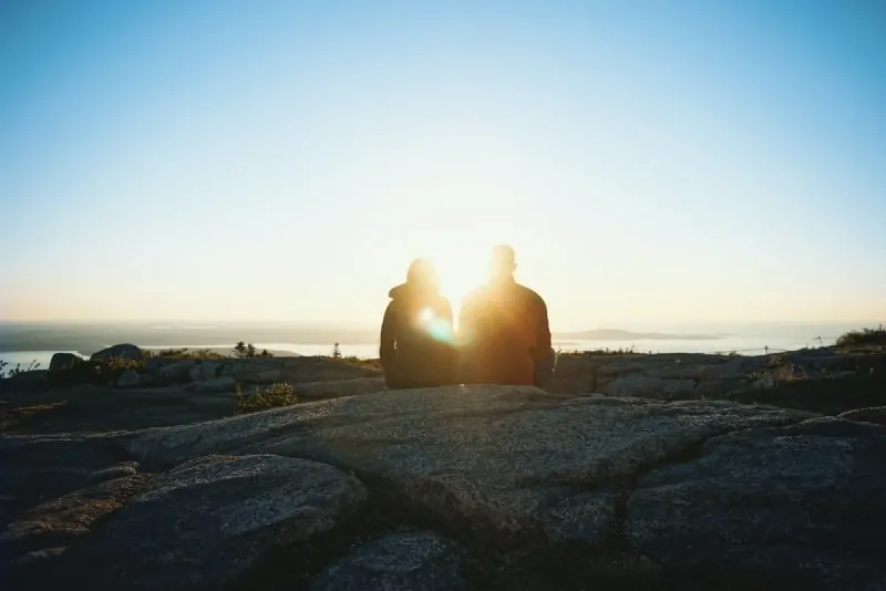 man and woman sitting on rock looking at sunrise